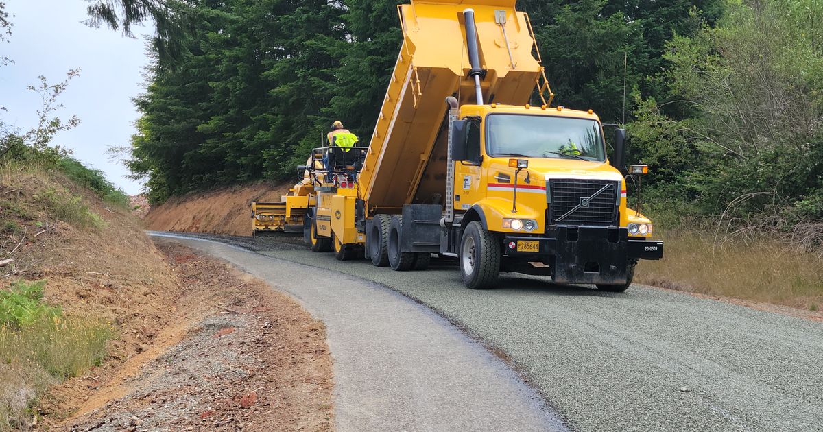 A yellow dump truck working on a road lined with trees.