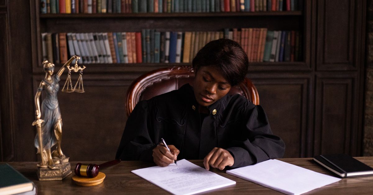 A judge in a black robe signs papers at a desk with a gavel and a statue of Lady Justice.