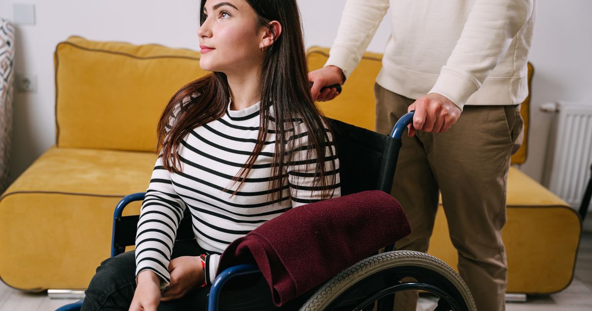 A young woman in a wheelchair at home with a caregiver standing behind her