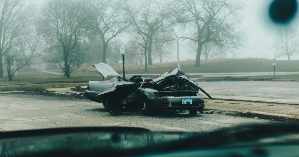 A wrecked silver car on a foggy street seen through a windshield