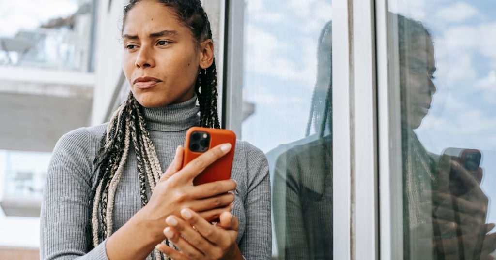 A woman holding a smartphone looks away with a worried expression beside a glass wall.